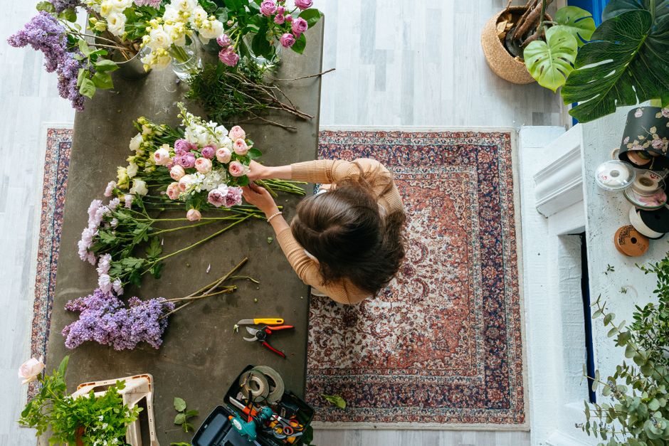 A florist arranging a bouquet of fresh flowers on a large grey worktable in a well-lit room. The bouquet includes pink roses, white carnations, and purple statice, with greenery interspersed. The flor