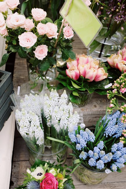 A variety of fresh flowers displayed on a wooden surface, including a bouquet of light pink roses with lush green leaves in a clear glass vase, and several arrangements of grape hyacinths in shades of
