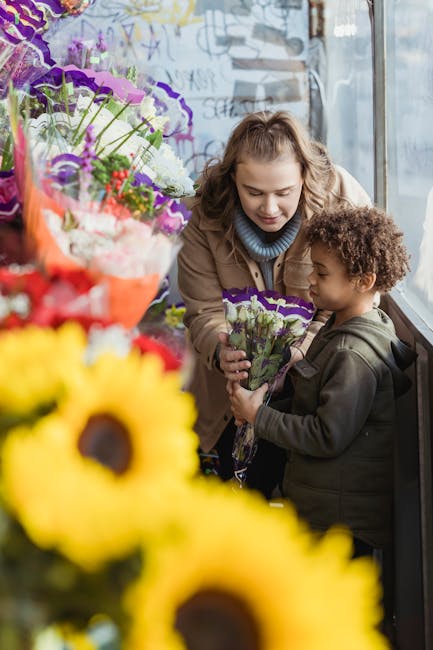 A young woman with light brown, curly hair and a child with dark, curly hair are inside a flower shop near a large window. The woman is smiling and dressed in a tan coat with a blue sweater underneath