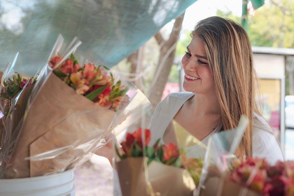 A young woman with long, light brown hair smiling warmly as she admires a bouquet of fresh flowers, including pink, peach, and red blooms, wrapped in brown kraft paper and transparent cellophane. She 